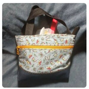 May include: A small, blue tote bag with a zipper closure and a patterned fabric lining. The bag has two black handles and is shown with various items inside, including a red pen, a white plastic container, and a blue and white card.