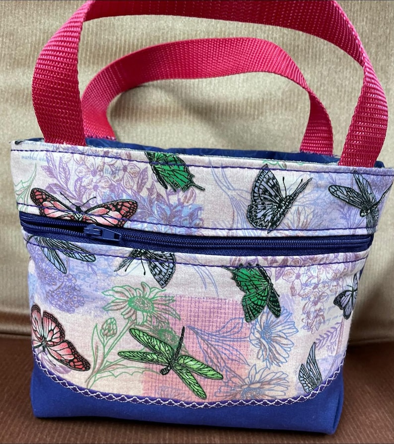 May include: A small, purple and pink tote bag with a zipper closure and two pink handles. The bag is decorated with a floral pattern featuring butterflies and dragonflies.