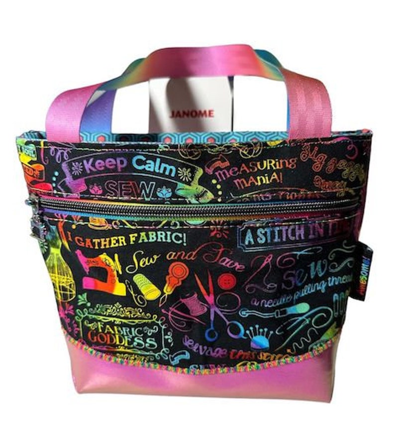 May include: A black fabric tote bag with a rainbow-colored design featuring sewing-themed text and illustrations. The bag has a zippered pocket and a pink iridescent bottom.