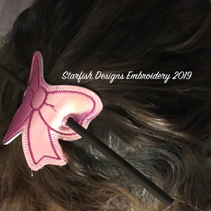 May include: A pink fabric bow with a black stick hair clip. The bow is decorated with a white stitch pattern. The text "Starfish Designs Embroidery 2019" is visible in the image.