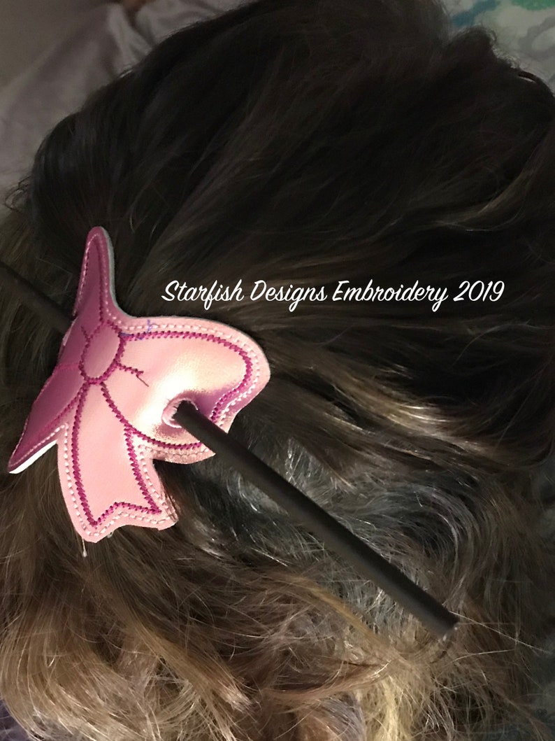 May include: A pink fabric bow with a black stick hair clip. The bow is decorated with a white stitch pattern. The text "Starfish Designs Embroidery 2019" is visible in the image.