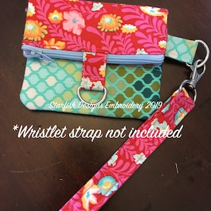 Può includere: Una piccola borsa in tessuto rosso e verde con una chiusura a zip e un anello di metallo. La borsa ha un motivo floreale e un motivo geometrico blu e verde. Il testo "Starfish Designs Embroidery 2019" è visibile sulla borsa. Il testo "*Wristlet strap not included" è visibile sotto la borsa.