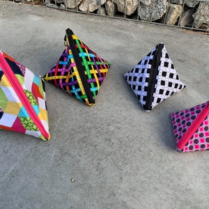 May include: Four pyramid-shaped zippered pouches with different colorful geometric patterns. The pouches are made of fabric and have pink zippers.