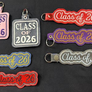 Puede incluir: Varios llaveros bordados con los textos "Class of 2026" y "Class of '26". Los llaveros son de varios colores, incluyendo rosa, gris, rojo, morado y azul. Cada llavero tiene un anillo metálico para sujetar llaves.