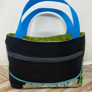 May include: A small black tote bag with a zippered pocket and a blue handle. The bag has a floral pattern on the bottom and a green lining.
