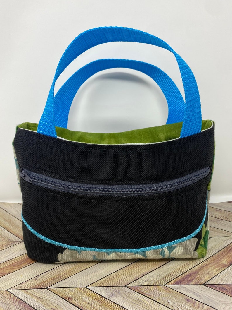 May include: A small black tote bag with a zippered pocket and a blue handle. The bag has a floral pattern on the bottom and a green lining.