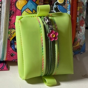 Pode incluir: Uma pequena bolsa de zíper verde limão com um amuleto de flor rosa. A bolsa tem um zíper verde e costuras coloridas ao redor das bordas.