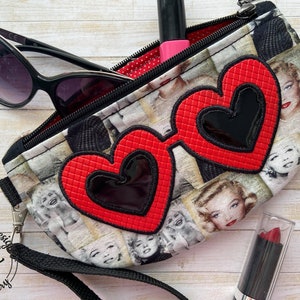 Può includere: Una pochette con cerniera a forma di occhiali da sole a cuore rosso con un cinturino nero e una stampa in bianco e nero del viso di Marilyn Monroe. La pochette è aperta e contiene un paio di occhiali da sole neri e un rossetto rosso.