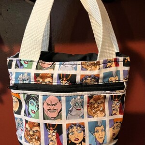 May include: A black and white tote bag with a zippered pocket. The bag features a repeating pattern of Disney villains' faces, including Ursula, Maleficent, and Cruella de Vil.
