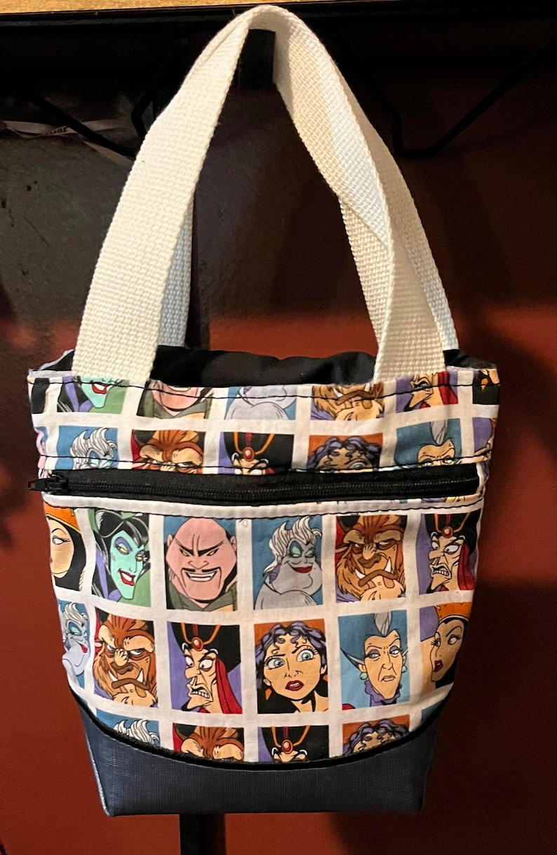May include: A black and white tote bag with a zippered pocket. The bag features a repeating pattern of Disney villains' faces, including Ursula, Maleficent, and Cruella de Vil.