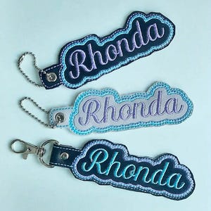 May include: Three personalized keychains with the name "Rhonda" embroidered in a cursive font. Each keychain has a different color scheme: dark blue with teal, light gray with blue, and dark blue with turquoise. They have a cloud-like shape.