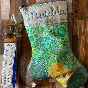 Könnte beinhalten: Ein weißer Strumpf mit einem grünen und blauen Kreis-Muster und dem Namen "Matilda" bestickt auf der Oberseite. Der Strumpf hat einen Reißverschluss und eine glitzernde grüne Spitze. Ein weißer und blauer Schlüsselanhänger mit einem goldenen Ring ist ebenfalls abgebildet.