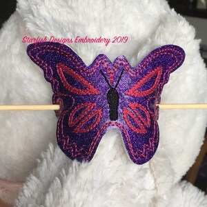 May include: Purple and red embroidered butterfly with glitter accents. The butterfly is attached to a white fuzzy surface with a wooden stick.