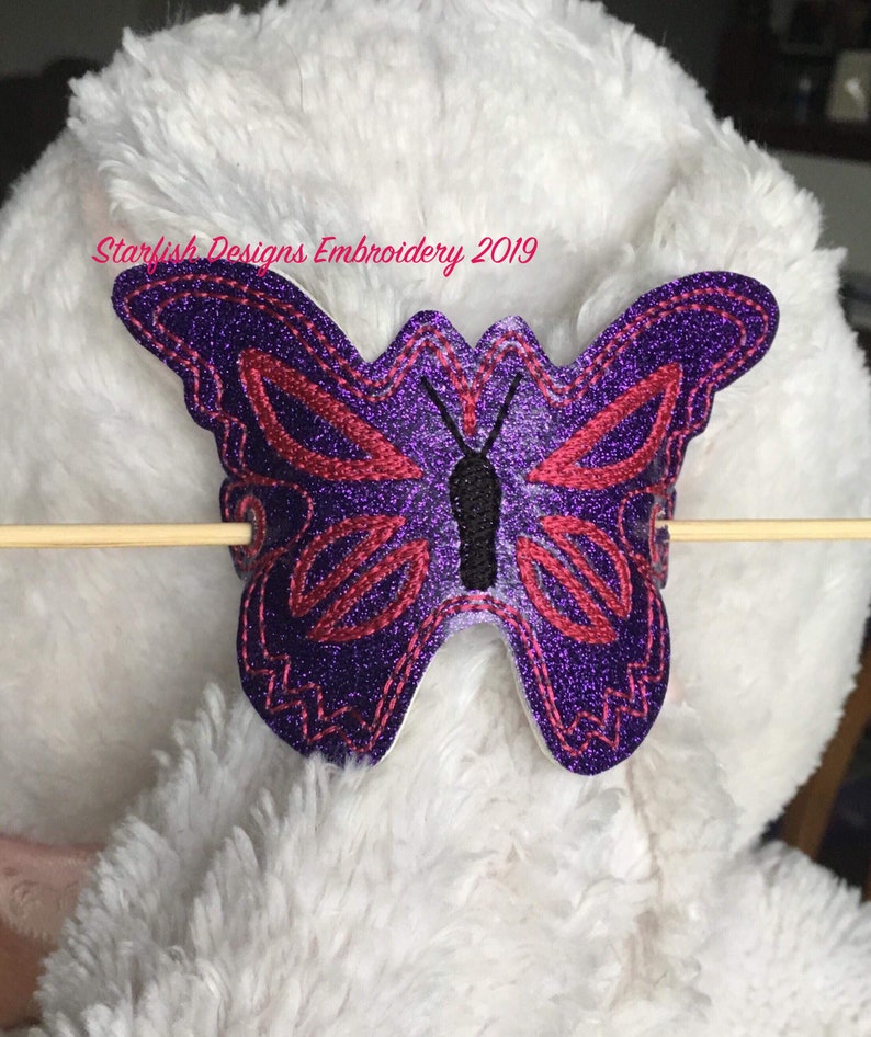 May include: Purple and red embroidered butterfly with glitter accents. The butterfly is attached to a white fuzzy surface with a wooden stick.
