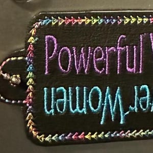 May include: Black leather tag with a colorful stitched border. The tag has the text "Powerful Women Empower" embroidered in purple and blue thread.