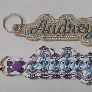 May include: Two embroidered keychains featuring the name Audrey in cursive script. The top keychain is tan cork with blue stitching and a silver ring. The bottom keychain has a black and white diamond pattern, purple snap, and rose gold clasp.