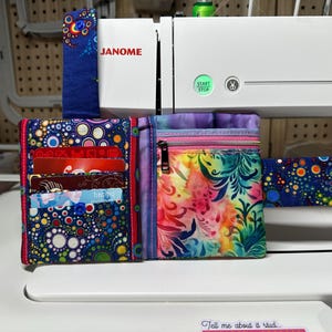 May include: A handmade fabric wallet and matching accessories. The wallet features a colorful design with a zipper pocket and card slots. The accessories have a similar pattern. The sewing machine in the background is a Janome.