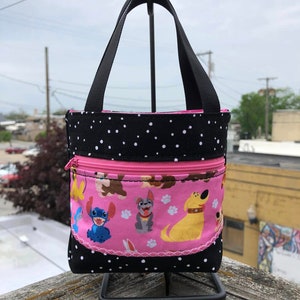May include: A small black and white polka dot tote bag with a pink zippered pocket. The pocket features a pink background with paw prints and cartoon images of Stitch and dogs from Disney's 'Lady and the Tramp'.
