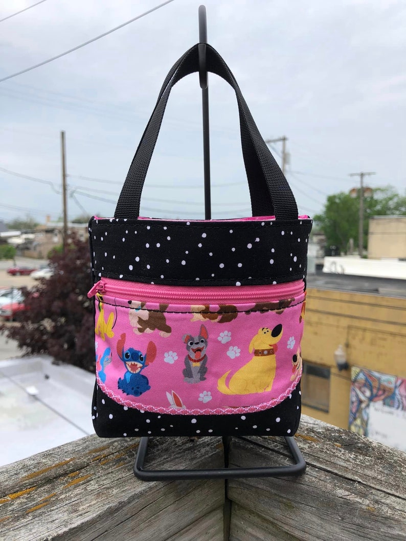 May include: A small black and white polka dot tote bag with a pink zippered pocket. The pocket features a pink background with paw prints and cartoon images of Stitch and dogs from Disney's 'Lady and the Tramp'.