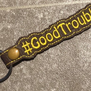 May include: Brown leather keychain with a metal ring and a gold button. The keychain has yellow embroidery that reads "#GoodTrouble".