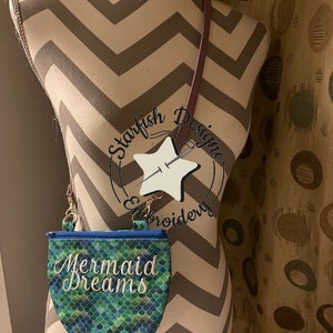 May include: A small, green and blue mermaid-themed bag with the text "Mermaid Dreams" embroidered on the front. The bag is hanging from a brown strap on a mannequin. The text "Daisy Mae Bag November 2019" is printed below the bag.