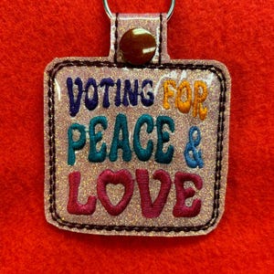 以下が含まれることがあります： レインボーカラーの「Voting for Peace & Love」という文字が書かれたピンクのグリッターキーホルダー。キーホルダーは「Love」の文字にハートの形をしています。