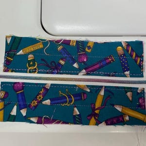 May include: A teal fabric pencil case with a colorful pencil pattern. The case is partially assembled, with a rainbow zipper and small fabric squares at each end. The text "Tell me about it said" is visible.