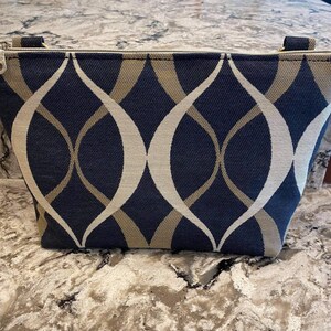 Può includere: Una pochette con cerniera nera e dorata con un motivo geometrico. La pochette è realizzata in tessuto e ha una chiusura a cerniera.