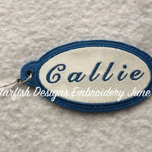 Puede incluir: Un llavero ovalado azul con fondo blanco y el nombre "Callie" bordado en hilo blanco.  Starfish Designs Embroidery June 2019 está impreso debajo del nombre.