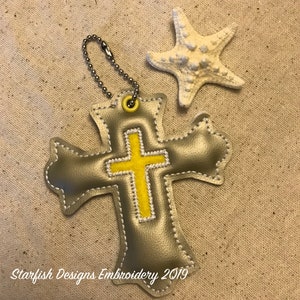 May include: A silver cross with a yellow embroidered cross on top. The cross is attached to a silver chain and has a small ring at the top. The cross is on a beige fabric background.