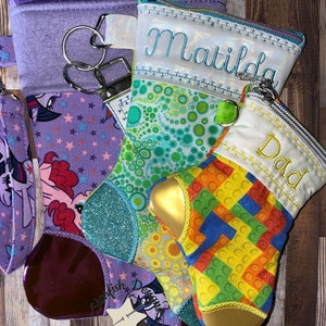 May include: Three colorful zippered pouches with embroidered names: Matilda, Dad, and a purple pouch with a My Little Pony design. The pouches are made of fabric and have a gold zipper pull.