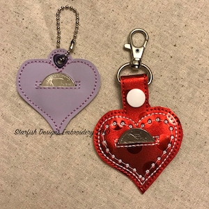 May include: Two heart-shaped coin purses with a silver chain and a silver clasp. The purse on the left is lavender with a silver coin inside. The purse on the right is red with a silver coin inside.