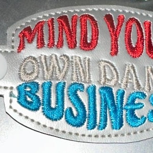 Könnte beinhalten: Ein silberner Schlüsselanhänger mit einem weißen ovalen Anhänger, auf dem "MIND YOUR OWN DAMN BUSINESS" in roter, weißer und blauer Stickerei steht.