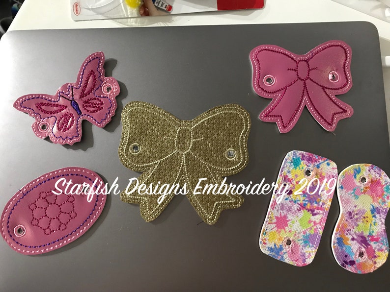 May include: Five embroidered patches with various designs, including a butterfly, two bows, a floral pattern, and two paint splattered patches. The patches are all pink, gold, and white with a white background.