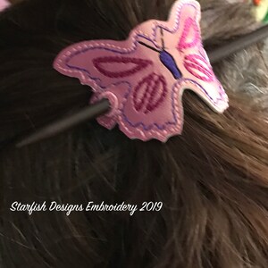 May include: A pink butterfly hair clip with purple and blue embroidery. The clip is attached to a dark brown stick. The text "Starfish Designs Embroidery 2019" is visible in the bottom left corner of the image.