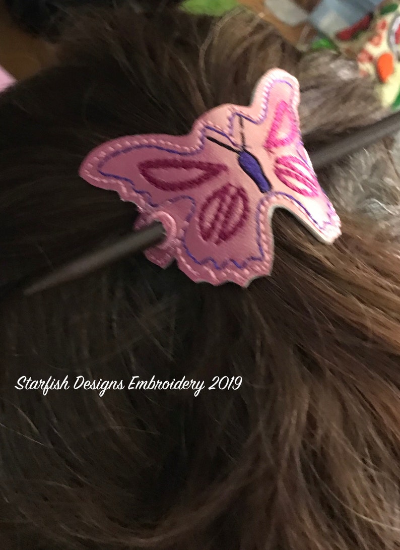 May include: A pink butterfly hair clip with purple and blue embroidery. The clip is attached to a dark brown stick. The text "Starfish Designs Embroidery 2019" is visible in the bottom left corner of the image.