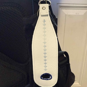 May include: White leather keychain with blue stitching and a blue oval design at the bottom. The keychain is attached to a silver metal clasp and is hanging on a black backpack.