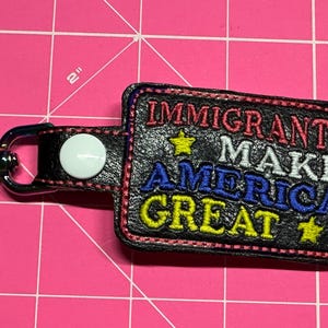 May include: Black rectangular keychain with the embroidered text "IMMIGRANTS MAKE AMERICA GREAT" in red, white, blue, and yellow. The keychain has a silver clasp and a black strap with a white button.
