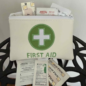 May include: A cream-colored first aid kit with a green cross emblem and the words "FIRST AID" embroidered in green. The kit is filled with bandages, antiseptic towelettes, and hand sanitizer packets. The kit is sitting on a black metal surface.