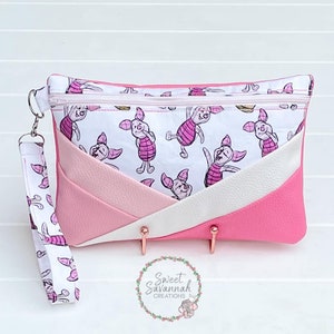 May include: Pink and white zippered pouch with a white strap. The pouch has a white background with a pink pig pattern. The pouch is made of faux leather and has a geometric design.