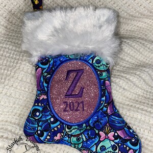 May include: A blue and purple Christmas stocking with a white faux fur cuff. The stocking features a glittery purple circle with the letter "Z" and the year "2021" embroidered in white. The stocking is decorated with a blue and purple pattern of cartoon characters.