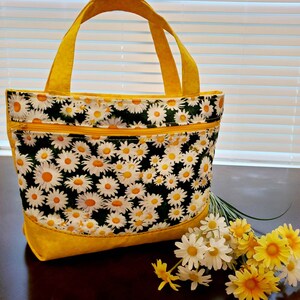 May include: A yellow tote bag with a daisy print and a yellow zipper. The bag has two handles and is sitting on a wooden surface with a bouquet of white and yellow daisies.