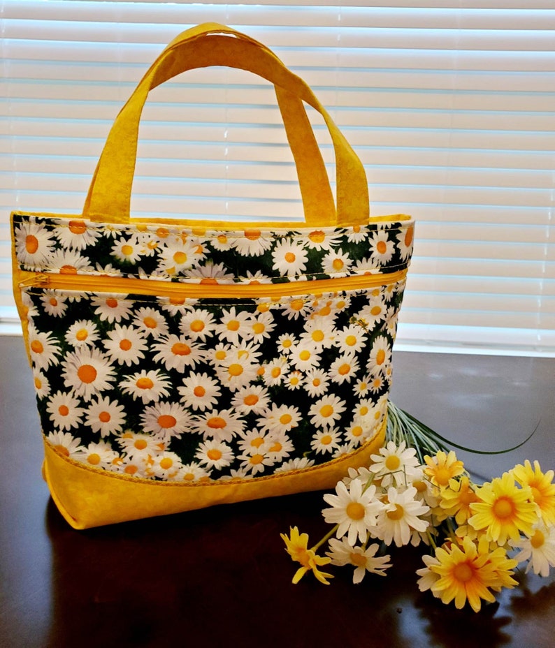 May include: A yellow tote bag with a daisy print and a yellow zipper. The bag has two handles and is sitting on a wooden surface with a bouquet of white and yellow daisies.