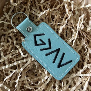 May include: A light blue leather keychain with the embroidered letters GNV in black. The keychain has a silver metal ring and a white stitched border. The background is filled with brown paper shreds.