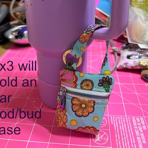 Può includere: Una piccola custodia per auricolari fantasia con chiusura a zip e manico, appesa a un bicchiere viola. La custodia presenta un motivo floreale nei toni del blu, rosa e arancione. Il testo sull'immagine recita "4x3 will hold an ear pod/bud case."