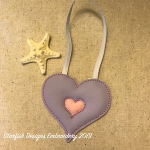 May include: A purple felt heart with a pink felt heart inside. The heart has a white ribbon loop for hanging. The heart is on a beige fabric background.  Starfish Designs Embroidery 2019.