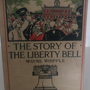 May include: Vintage book cover titled "The Story of the Liberty Bell" by Wayne Whipple. The cover features an illustration of a crowd of people in front of a building and a drawing of the Liberty Bell. The book is displayed on a metal stand.