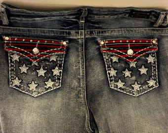 Pode incluir: Calções de ganga azul com dois bolsos traseiros com fecho de aba. Os bolsos são decorados com um padrão de riscas vermelhas e azul-marinho, tachas prateadas e uma estampa de estrelas brancas. O tecido tem uma lavagem desbotada e botões.