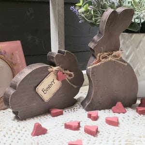 May include: Two wooden bunny figurines with a distressed gray finish, adorned with twine bows and small pink hearts. One bunny has a tag that says "Bunny". Scattered around are small, pink heart-shaped decorations, creating a rustic Easter theme.