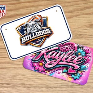 Puede incluir: Dos placas de matrícula. Una placa blanca con borde azul presenta un logotipo de bulldog y la palabra "BULLDOGS". La otra placa es rosa con el nombre "Kaylee" en cursiva, rodeado de flores y corazones.